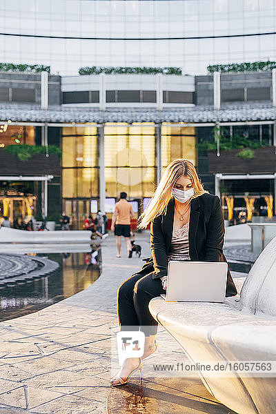 Geschäftsfrau mit Maske  die einen Laptop benutzt  während sie auf einem Sitz vor einem Gebäude sitzt