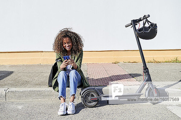 Glückliche junge Frau  die ihr Smartphone benutzt  während sie auf dem Bürgersteig sitzt und einen Elektroroller fährt