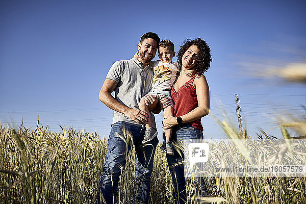 Smiling parents carrying son while standing amidst crops against clear blue sky