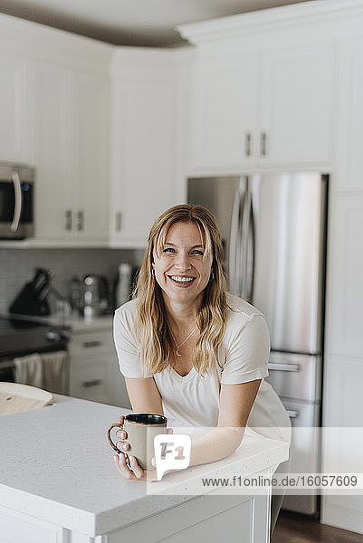 Lächelnde Frau  die eine Tasse auf dem Küchentisch hält  während sie zu Hause steht
