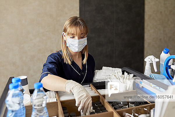 Zimmermädchen mit Maske ordnet Flaschen auf einem Rollwagen an der Wand im Hotelflur