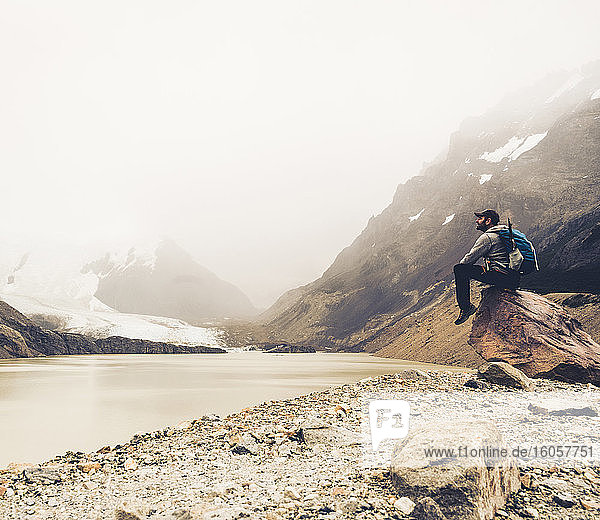 Älterer Mann mit Rucksack  der auf einem Felsen sitzend den See betrachtet  Patagonien  Argentinien