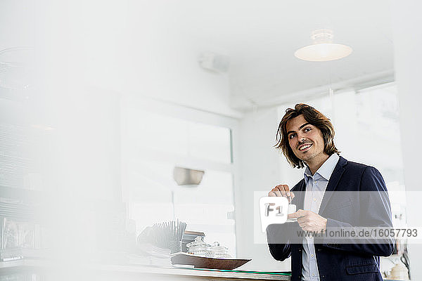 Lächelnder männlicher Unternehmer schaut weg  während er eine Kaffeetasse in einem Café hält