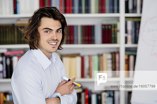 Smiling businessman with arms crossed sitting against bookshelf in office