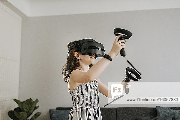 Girl wearing virtual reality simulator playing with joysticks at home