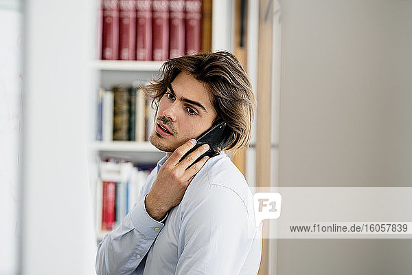 Männlicher Berufstätiger  der über sein Smartphone spricht und wegschaut  während er im Büro sitzt
