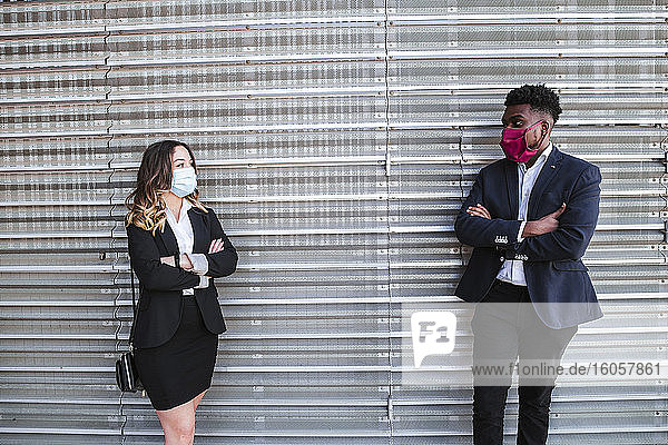 Business professionals wearing masks with arms crossed talking while standing against wall