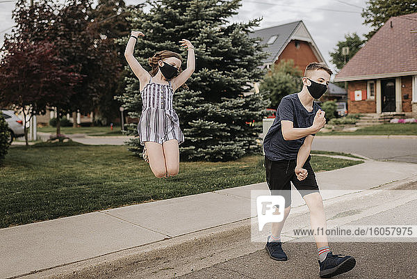 Unbekümmerte Geschwister mit Masken tanzen und springen auf der Straße in der Stadt