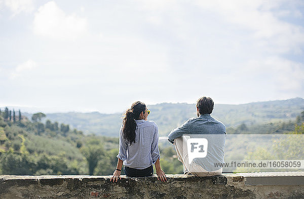 Couple looking at view while sitting on retaining wall against sky during sunny day