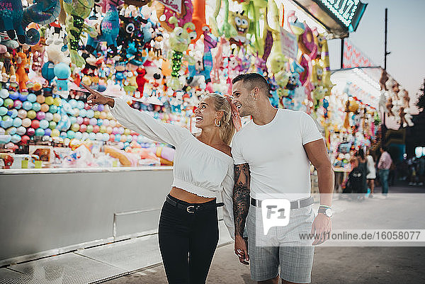 Happy woman pointing at stall while standing with boyfriend in amusement park