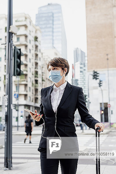 Geschäftsfrau mit Maske  die ein Smartphone hält  während sie mit einem Koffer in der Stadt steht