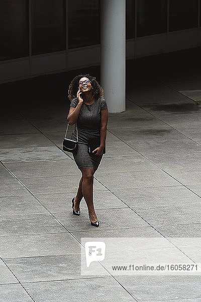 Fröhliche junge Geschäftsfrau  die über ihr Smartphone spricht  während sie auf einem Fußweg in der Stadt spazieren geht