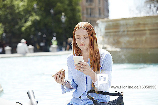 Frau  die ein Smartphone benutzt  während sie an einem Brunnen in der Stadt einen Snack zu sich nimmt