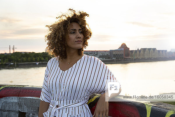 Nachdenkliche Frau mit lockigem Haar steht am Geländer gegen den Fluss in der Stadt bei Sonnenuntergang