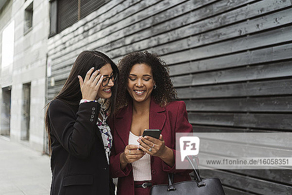 Lächelnde junge multiethnische Geschäftskollegen  die sich ein Smartphone an einer Wand in der Stadt teilen