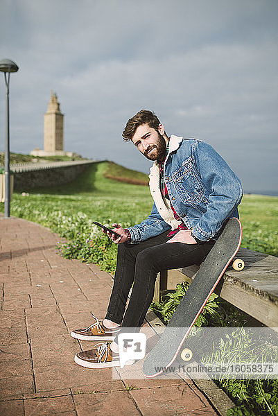 Gut aussehender junger Mann  der sein Smartphone benutzt  während er mit einem Skateboard auf dem Sitz sitzt
