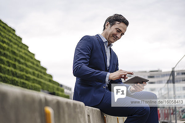 Mature businessman sitting on a wall in the city using tablet