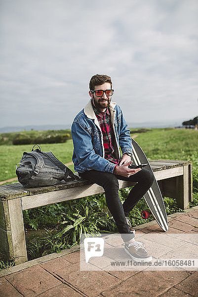 Gut aussehender Mann mit Sonnenbrille sitzt mit Skateboard auf dem Sitz gegen den bewölkten Himmel