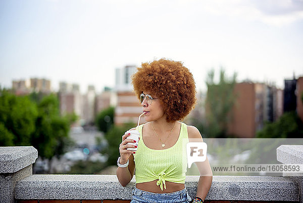 Mid adult woman with afro hair drinking juice while standing against sky in city
