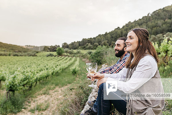 Glückliches Paar mit Weißweingläsern in der Hand auf einem Weinberg sitzend