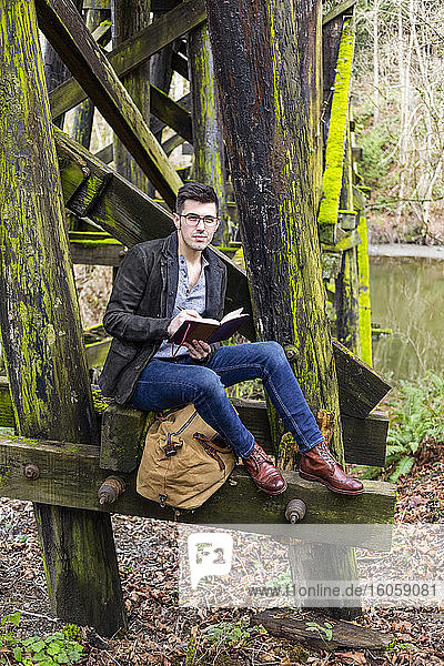 Young man in sitting on a moss-covered trestle bridge beside a pond reading from a book in a quiet location; Bothell  Washington  United States of America