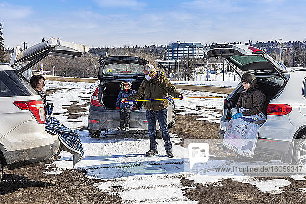 Familien sitzen hinten in ihren Fahrzeugen auf einem Parkplatz  den sie während der Covid-19-Weltpandemie besuchen  ein Mann misst ihren Sicherheitsabstand voneinander mit einem Maßband; St. Albert  Alberta  Kanada