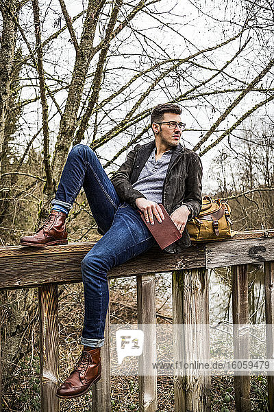 Porträt eines jungen Mannes im Wald  der auf einem Zaun sitzt und sich mit einem Buch in der Hand zurücklehnt; Bothell  Washington  Vereinigte Staaten von Amerika