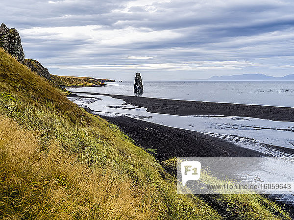 Hohe Felsformation und grasbewachsene Hänge entlang der Uferlinie eines Fjordes; Island