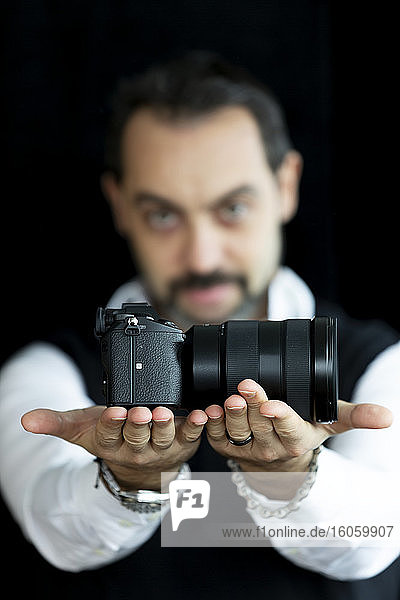 Portrait of a male photographer standing with his camera held out in focus against a black background; Studio