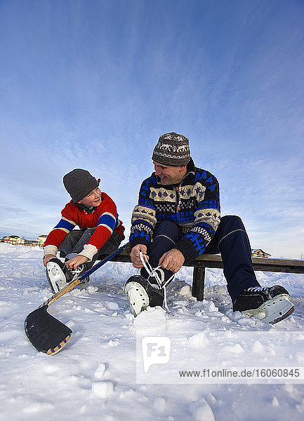 Ein Vater und sein Sohn schnüren ihre Schlittschuhe auf einer Bank auf einem zugefrorenen See für ein Hockeyspiel im Freien; Alberta  Kanada