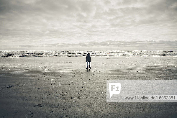 Teenager Junge steht auf riesigen Strandwellen und bedecktem Himmel in der Ferne