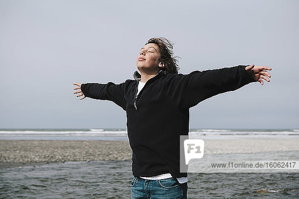 Teenage boy on beach with arms outstretched towards breeze ocean in distance
