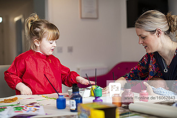 Young girl using paints at kitchen table with mother sitting nearby holding baby
