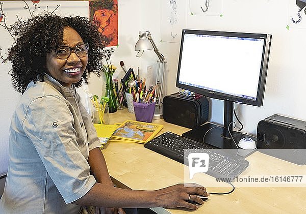 Dunkelhäutige  lächelnde Frau mit Brille arbeitet am Schreibtisch vor Computer  Home Office  Blick in die Kamera  Deutschland  Europa