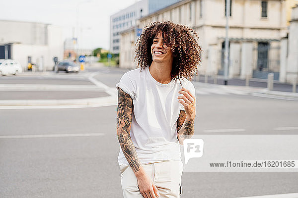Lächelnder Mann mit tätowierten Armen und langen braunen lockigen Haaren steht auf einer Straße.