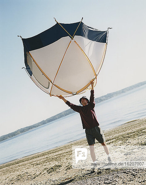 Ein Mann  der ein kleines Kuppelzelt über seinem Kopf hält und am Strand steht
