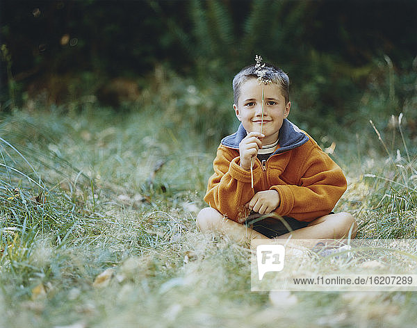 A young boy sits in field of tall grass  holding blade of grass