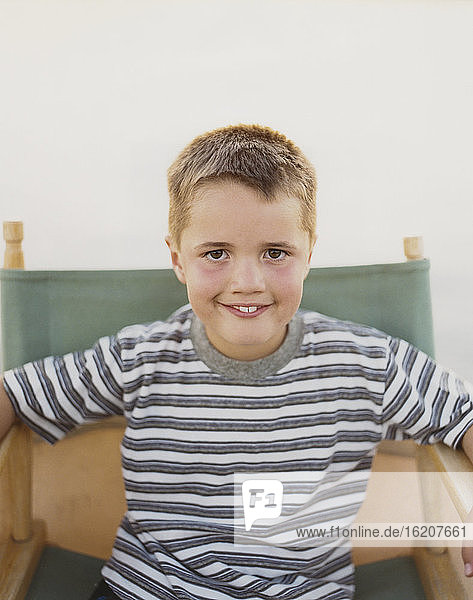 Ein selbstbewusster Junge sitzt auf einem klappbaren Regiestuhl und lächelt mit einem zahnigen Lächeln.