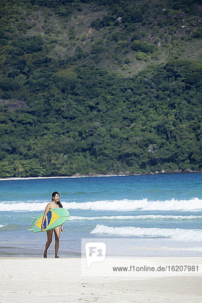 Strandaufnahme eines japanischen Brasilianers (Nipo-brasileiro) im Bikini  der ein mit der brasilianischen Flagge geschmücktes Surfbrett trägt  Brasilien  Südamerika
