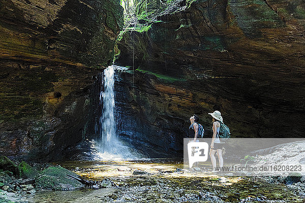 Zwei junge Wanderer beim Canyoning einen Fluss hinauf und stehen in einer Höhle im Regenwald vor einem unberührten Wasserfall  Minas Gerais  Brasilien  Südamerika