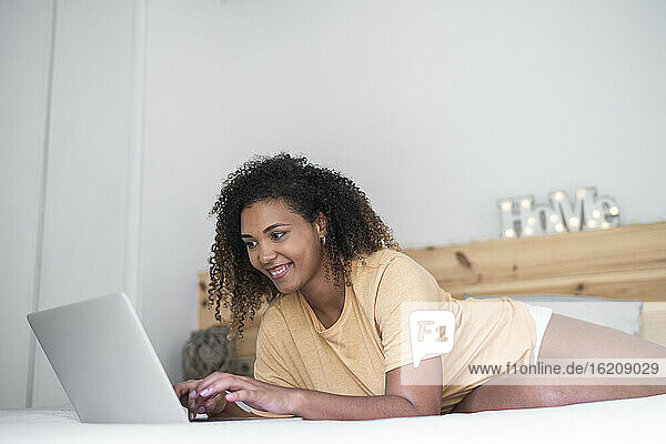Smiling young woman with curly hair using laptop while lying on bed at home