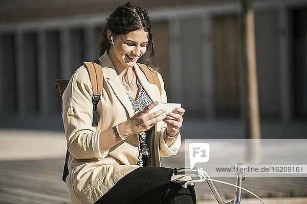 Junge Frau  die ihr Smartphone benutzt  während sie auf einem Fahrrad in der Stadt bei Sonnenschein sitzt