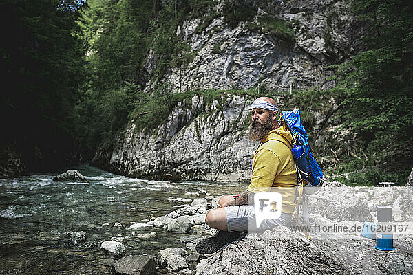 Hiker with full beard and yellow hoodie sitting on stone next to river