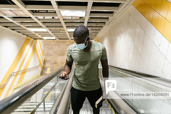 Junger Mann mit Maske benutzt sein Smartphone  während er auf einer Rolltreppe in einer U-Bahn-Station steht