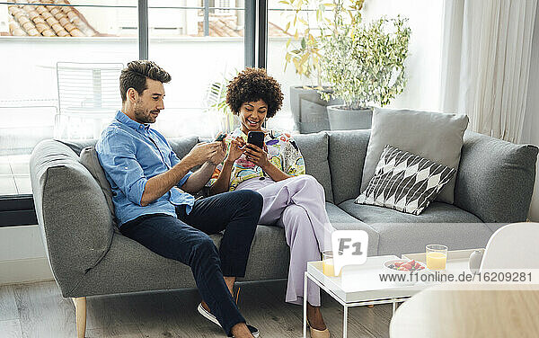 Multiethnisches Paar benutzt Smartphones  während es auf dem Sofa im Penthouse sitzt