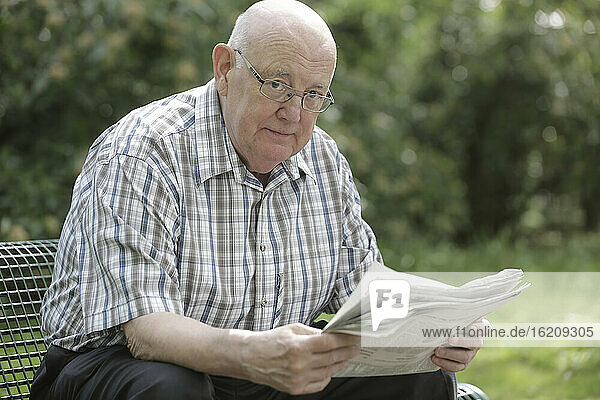 Germany  North Rhine Westphalia  Cologne  Portrait of senior man reading newspaper on bench in park