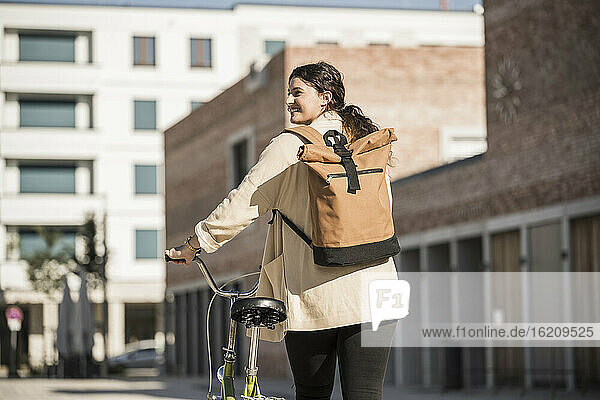 Young woman carrying backpack while walking with bicycle on city street