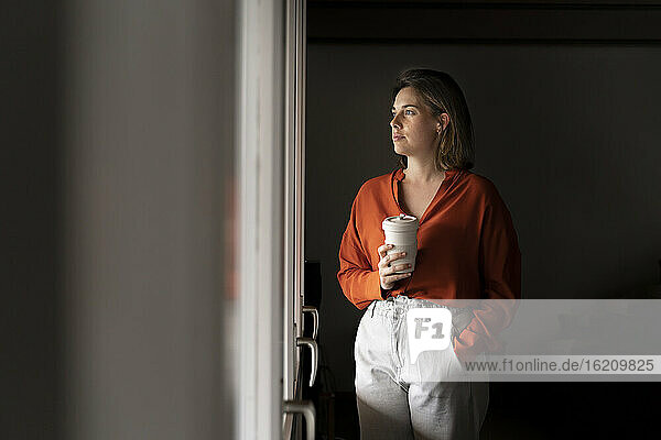 Businesswoman with coffee cup looking through window at home