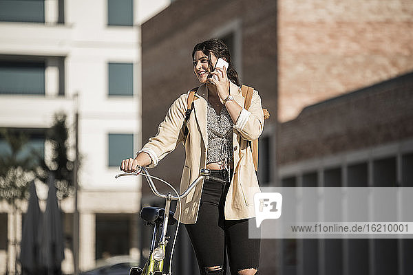 Smiling young woman talking over mobile phone while walking with bicycle in city