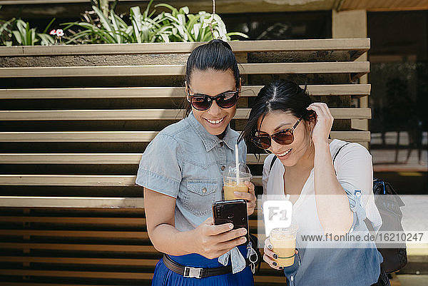 Freundinnen mit Sonnenbrille  die ein Foto über ein Smartphone in der Stadt betrachten